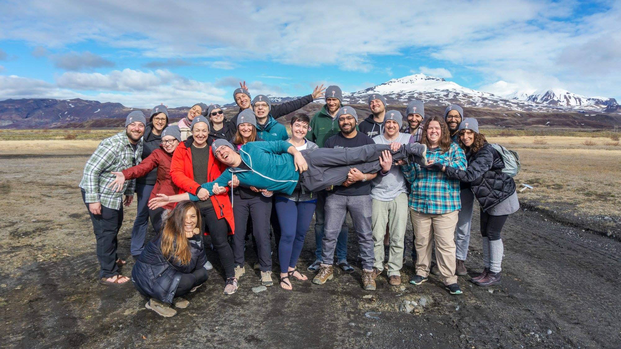 Group of friends posing together outdoors in Iceland, laughing as they hold one person horizontally, with rugged volcanic terrain and snow-capped mountains stretching across the background