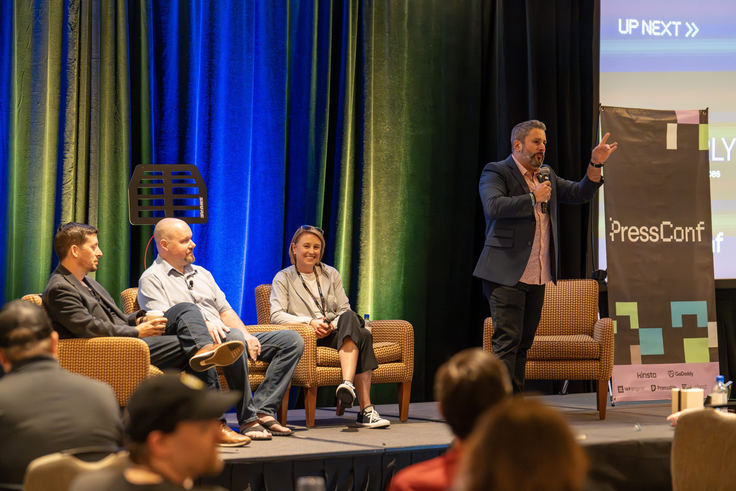 Panel discussion at PressConf with three seated speakers on stage while a host stands with a microphone, gesturing to the audience. Colorful stage lighting and a PressConf banner frame the scene.